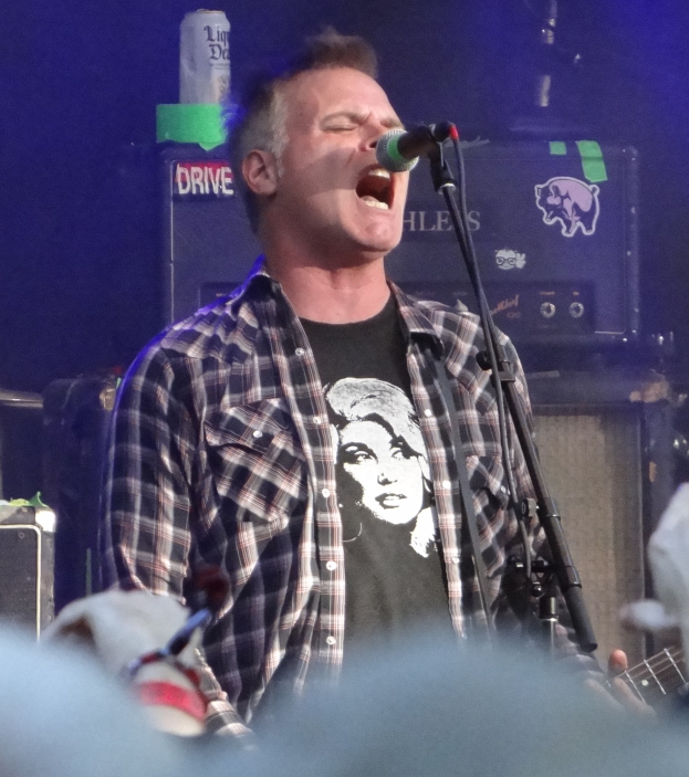 Ein Mann, der auf der Bühne in ein Mikrofon singt und eine Gitarre hält, mit einem unscharfen Gesicht im Vordergrund und verschiedenen Gegenständen im Hintergrund.