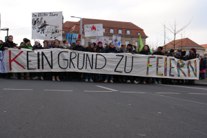 Demonstranten mit einem Transparent mit der Aufschrift "Kein Grund zu Feiern" gegen deutsche Sparmaßnahmen, im Hintergrund Straßeninfrastruktur und Gebäude bei klarem Himmel.
