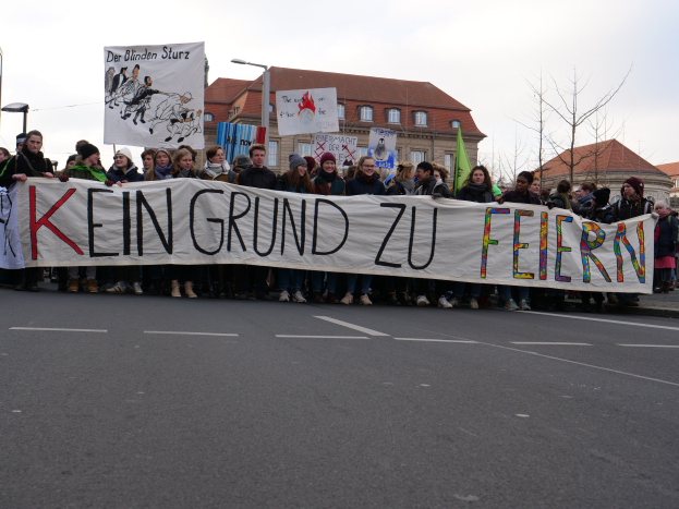 Demonstranten mit einem Transparent mit der Aufschrift "Kein Grund zu Feiern" gegen deutsche Sparmaßnahmen, im Hintergrund Straßeninfrastruktur und Gebäude bei klarem Himmel.