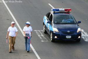 Zwei Frauen gehen auf der Straße, eine hält einen rosafarbenen Zettel und trägt eine rosafarbene und weiße Mütze, die andere hält einen rosafarbenen Zettel und trägt eine blaue und weiße Mütze, mit einem patrouillierenden Polizeiwagen und seinem Fahrer auf der rechten Seite.