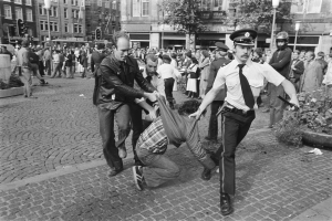 Ein Schwarz-Weiß-Bild eines Mannes, der von zwei Polizeibeamten auf der Straße weggezerrt wird, mit vielen Menschen, Bäumen, Gebäuden, Laternen, Verkehrsampeln und Topfpflanzen im Hintergrund und einem Mannenschuh im Vordergrund.