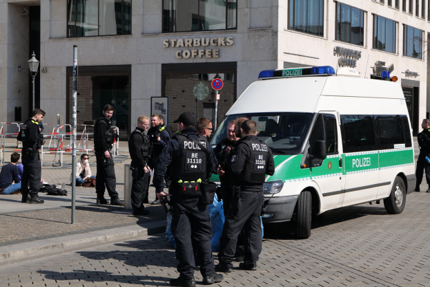 Eine Gruppe von Polizisten steht vor einem Starbucks-Café, rechts davon steht ein Van und links ein paar Menschen; im Hintergrund sind ein Gebäude, ein Schild, ein Laternenpfahl und ein Zaun zu sehen.