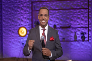 Stephen Colbert, Moderator von *The Daily Show with Trevor Noah*, steht auf einer Bühne und hält ein Mikrofon, trägt einen Anzug, Krawatte und Brille, mit einem Scheinwerfer und Ständer dahinter.