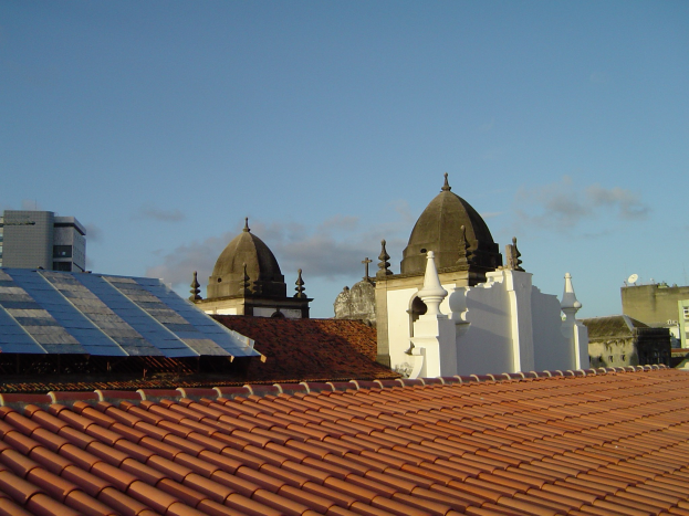 Stadtpanorama mit einigen Gebäuden im Vordergrund, einem blauen Himmel im Hintergrund und Solarpanelen auf einem Dach, die den Einsatz erneuerbarer Energien anzeigen.