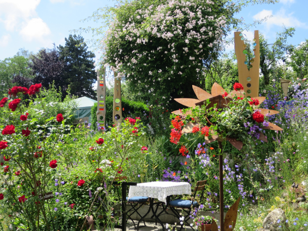 Ein Garten mit farbenfrohen Blumen und Pflanzen, der einen Tisch mit einer Decke und Stühlen zeigt, vor Bäumen und einem bewölkten Himmel auf der Chelsea Flower Show.