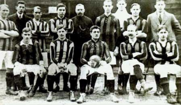 Schwarzes und weißes Foto einer Fußballmannschaft, einige stehen und einige sitzen, vor einer Wand mit dem Text 'Ballymena Football Club' unten.