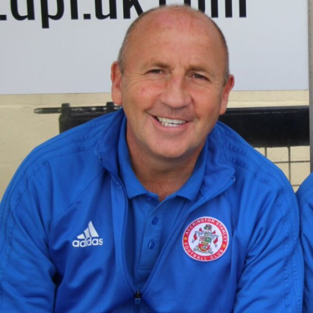 Zwei Männer in einem Raum, einer in einer blauen Jacke, der neben einem anderen Mann lächelt, mit einer Wandtafel im Hintergrund.