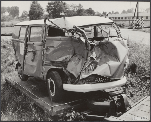 Schwarzes und weißes Foto eines schwerbeschädigten Autos mit eingedrückter Front und zersplitterten Fenstern auf einem Tieflader, umgeben von Gras und Pflanzen mit Bäumen und einem Gebäude im Hintergrund.