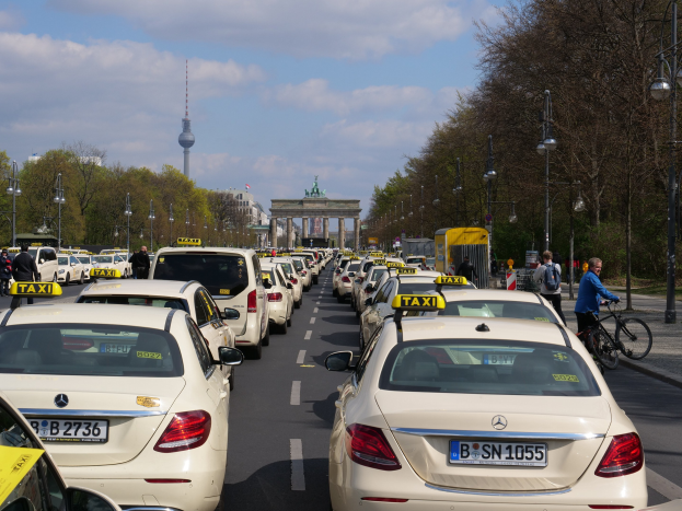 Eine lange Reihe von Taxis, die an einer belebten Straße in Berlin, Deutschland, geparkt sind, mit fahrenden Fahrzeugen, Radfahrern, Fußgängern, Bäumen, Laternenpfählen, Gebäuden, einem Bogen und einem Turm im Hintergrund unter einem bewölkten Himmel.