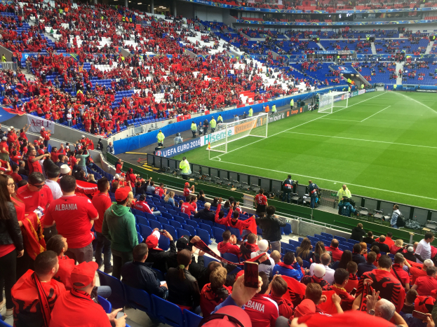 Ein volles Stadion mit Zuschauern, einige sitzen, andere stehen, bei einem Fussballspiel, mit Bannern, einem Torpfosten und Deckenleuchten.