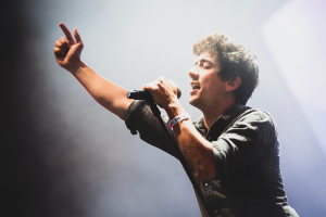 Ein Mann in schwarzem Hemd und Jeans singt leidenschaftlich in ein Mikrofon auf der B├╝hne, mit geschlossenen Augen und einem jubelnden Publikum im Hintergrund.