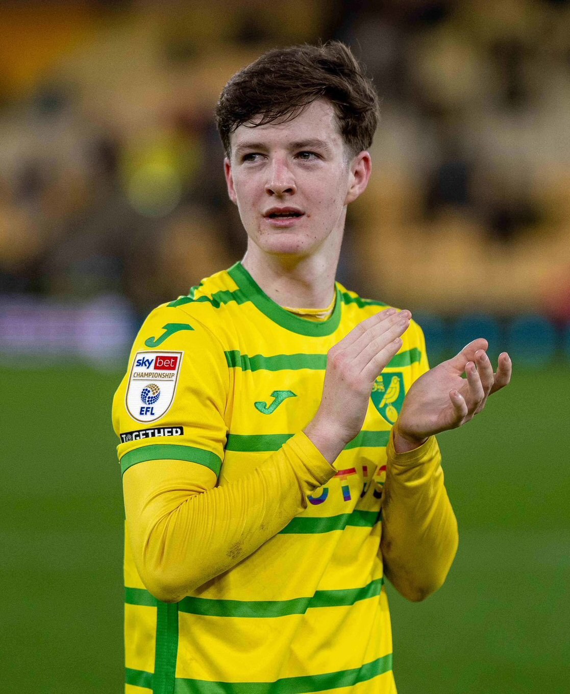 Ein junger Mann in einem gelben und grünen T-Shirt steht auf einem Fußballfeld und klatscht begeistert in die Hände vor einem unscharfen Hintergrund.
