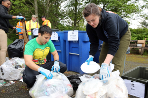 Zwei Personen in Handschuhen sammeln Müll auf Tellern in einem Park, umgeben von Müll, mit einem Müllcontainer und einer Bank rechts und Bäumen und Himmel im Hintergrund.