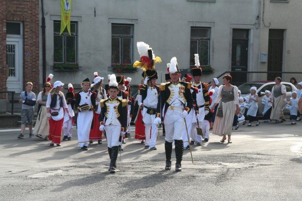 Eine Gruppe von Menschen geht auf der Straße in einer Parade, einige in Kostümen und mit Stöcken, mit Gebäuden, Pflanzen, einer Fahne und Fahrzeugen im Hintergrund.