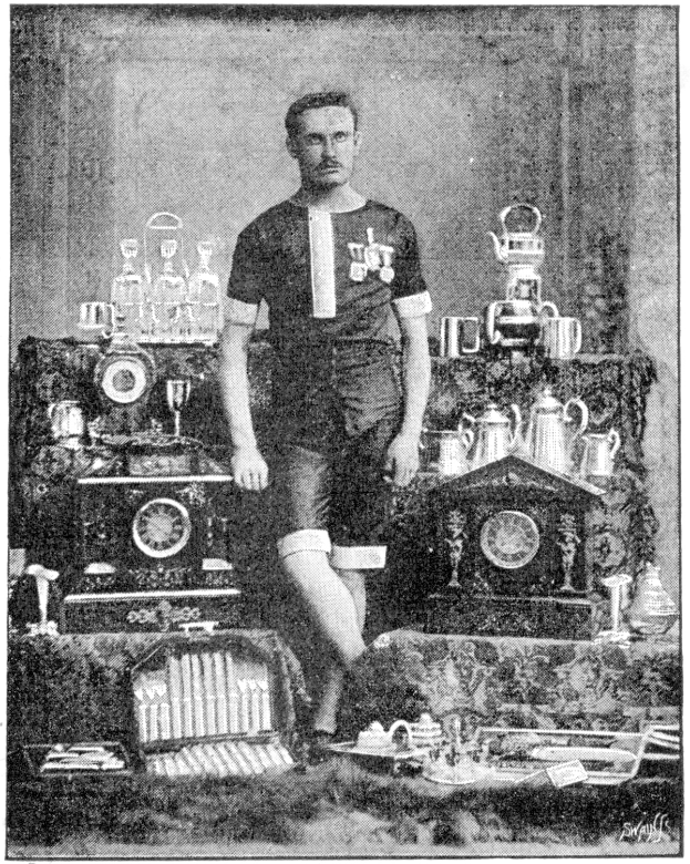 Schwarzes und weißes Foto eines Mannes in einem Fußballtrikot, der vor einem Tisch mit Pokale, Trophäen und anderen Gegenständen steht, beschriftet mit "1909-1910 WM-Sieger."