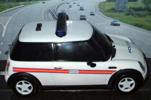 Ein weißer Polizeiwagen steht im Vordergrund mit mehreren Autos, die sich auf der Straße dahinter bewegen, umgeben von Gras und Bäumen auf beiden Seiten in einer ländlichen Umgebung.