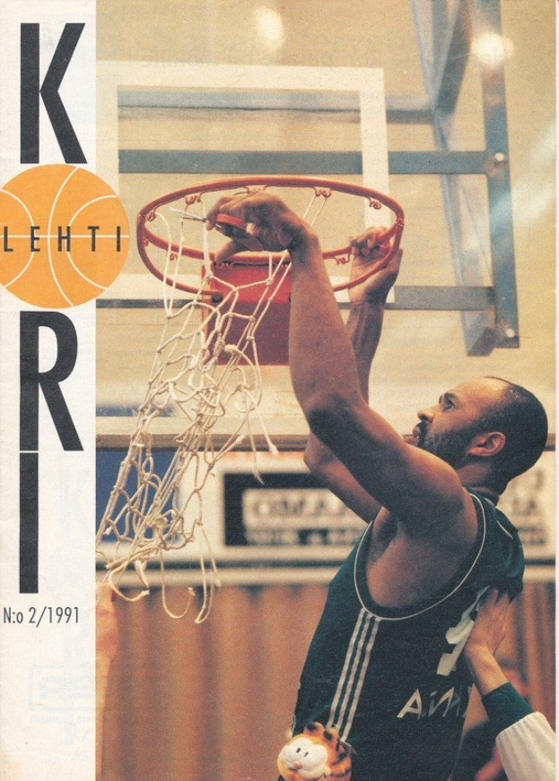 Ein Basketballspieler wirft den Ball in den Korb, während eine andere Person daneben steht, mit einem Basketballkorb und einer Wand im Hintergrund und einer Kobe-Bryant-Basketballkarte links sichtbar.