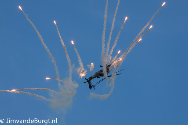Ein Hubschrauber fliegt durch die Luft mit einer Rauchspur, die eine Feuerspur am Himmel hinterlässt, mit Text in der unteren linken Ecke.