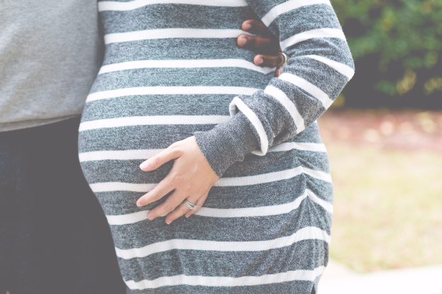 Schwangere Paare stehen im Freien mit Händen auf dem Bauch, tragen Schwangerschaftskleidung, umgeben von Bäumen.