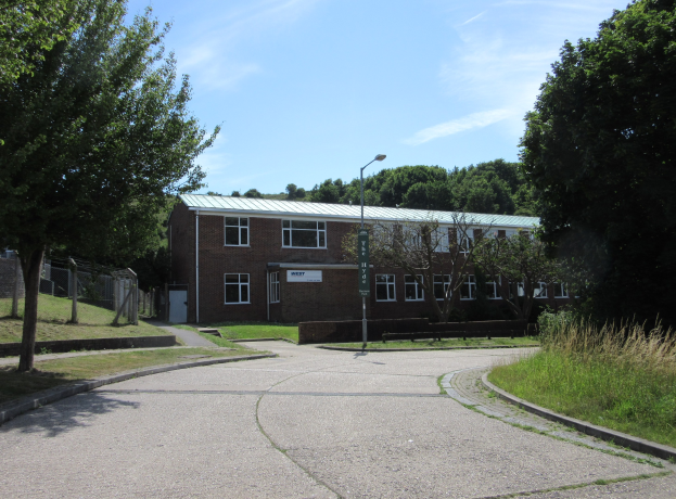 Ein Schulgebäude mit Fenstern, einer Straßenlaterne, einem Schild, Bäumen, Gras, einem Zaun und einem bewölkten Himmel, das sich in einem Park mit saftigem Gras befindet.