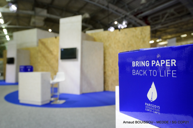Ein Messestand mit einem blauen und weißen Schild, auf dem "Bring Paper Back to Life" steht, auf der rechten Seite, mit unscharfen Stühlen, Tischen, einem wandmontierten Fernseher und Deckenleuchten im Hintergrund.