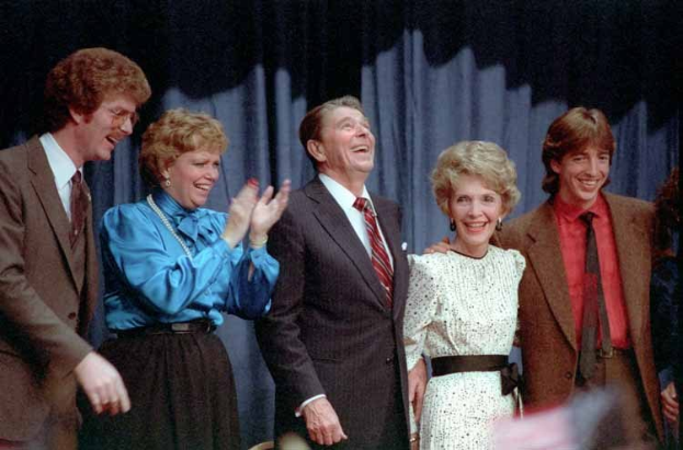 Group of people from *The Brady Bunch* standing on a stage, smiling and clapping with a curtain backdrop.