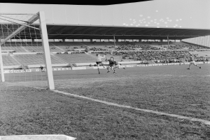 Ein Schwarz-Weiß-Foto eines Fußballspiels im Gange, mit Spielern auf dem Feld und Zuschauern im Hintergrund.