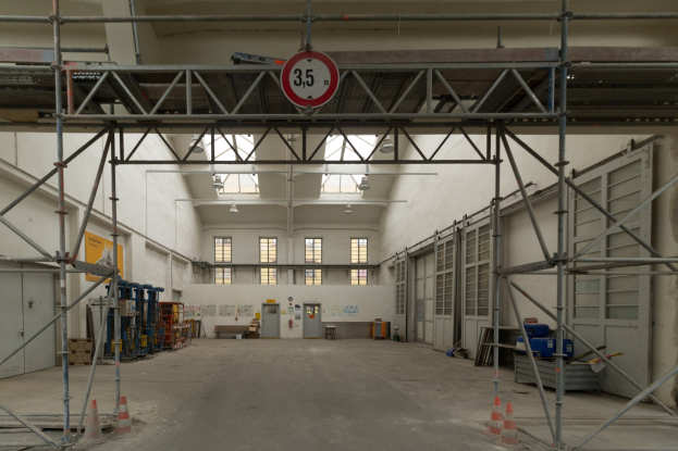 Lagerhalle mit ausgedehnten Gerüsten, Verkehrskegeln, Metallstangen, einem Schild, Maschinen, einer Tür, Fenstern, Wänden, Deckenlampen und einem Dach.