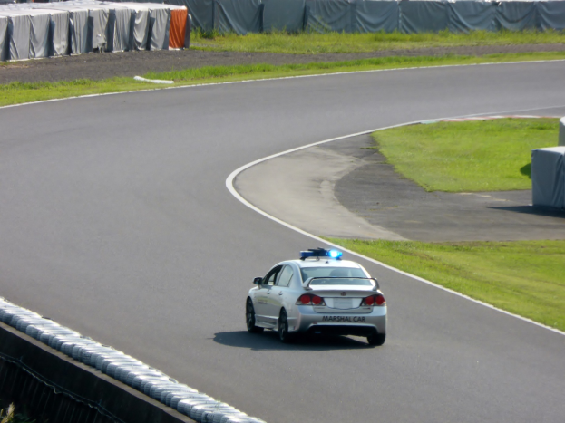 Polizeiauto fährt auf einer Rennstrecke mit Gras auf beiden Seiten, Menschen und Zelte sind im Hintergrund zu sehen.