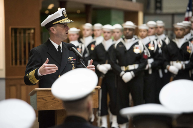 Ein Mann in militärischer Uniform spricht an einem Pult während einer Graduierungsfeier der US Naval Academy, mit Absolventen in weißen Mützen, bewaffneten Personen und einer Flagge im Hintergrund.
