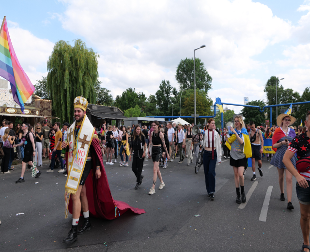 Eine Gruppe von Menschen marschiert bei der Pride Parade 2018 mit einer Regenbogenflagge und Musikinstrumenten, umgeben von Laternenmasten, Bäumen, Hütten und einem bewölkten Himmel.