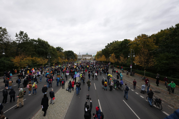 Eine große Gruppe von Menschen marschiert eine von Bäumen gesäumte Straße in Berlin entlang und hält Kameras und Schilder hoch, mit einem Gebäude und einem klaren Himmel im Hintergrund.