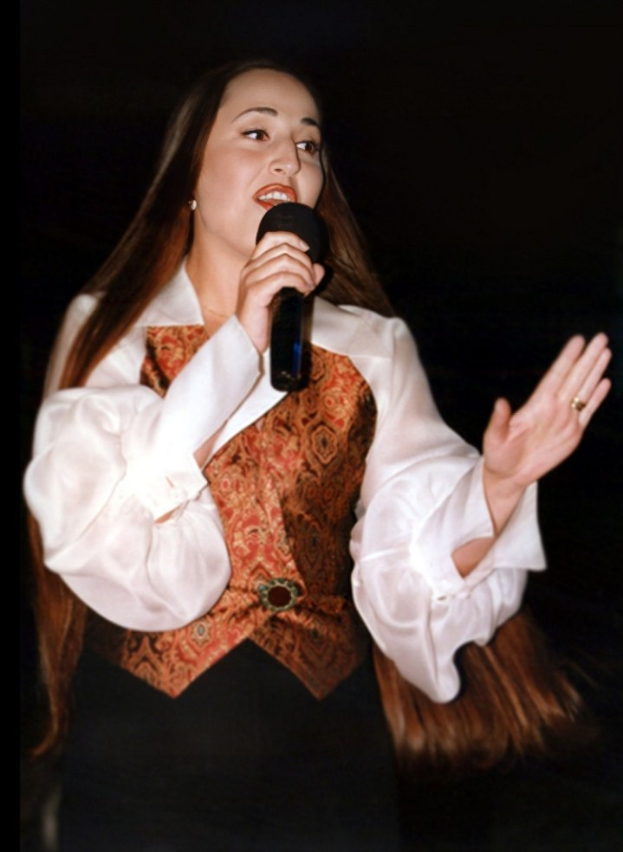 Eine Frau in einem weißen Hemd und einer braunen Weste singt in ein Mikrofon und strahlt dabei Selbstvertrauen aus, während sie es festhält und ihr Haar zu einem Dutt gebunden hat und ihren Blick auf das Publikum richtet.