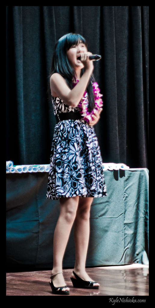 Eine Frau in einem bunten Kleid singt auf der B├╝hne in ein Mikrofon, mit einem Tisch und Papieren im Hintergrund hinter einem Vorhang.