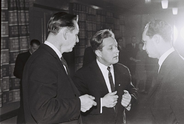 Drei Männer in Anzügen unterhalten sich in einem schwach beleuchteten Raum mit Bücherregalen im Hintergrund.