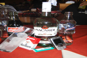 Eine Sammlung von Flaschen, darunter Alkohol, Wasser und eine Coca-Cola, ein Glas mit Eisw√ľrfeln, ein Feuerzeug und ein Mobiltelefon auf einem Tisch mit Stühlen und Menschen, die Wein im Hintergrund trinken.