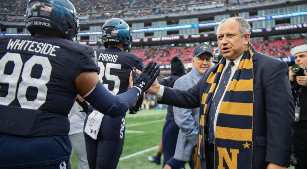 Ein Mann im Anzug und Krawatte gibt einem Fußballspieler die Hand auf einem Feld, umgeben von einer Gruppe von Menschen, mit einer Person, die eine Kamera auf der rechten Seite hält, und einem Stadion mit Zuschauern und Tafeln im Hintergrund, die "Notre Dame Football Coach Mike McCarthy und die Fighting Irish" lesen.