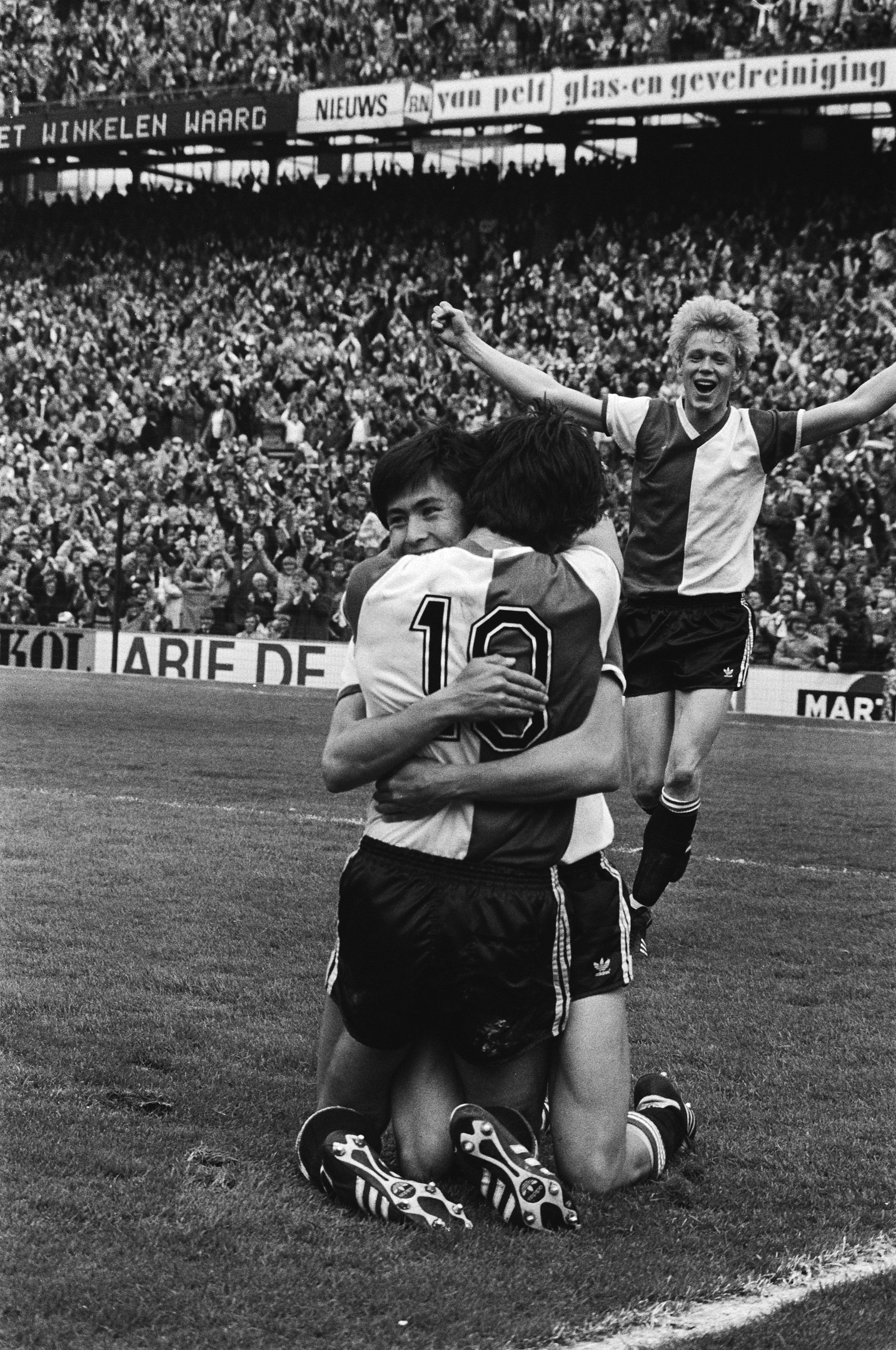 Drei Fußballspieler umarmen sich auf dem Boden nach einem Tor, mit jubelnder Menge und schwenkenden Fahnen im Hintergrund, in einem schwarz-weißen Stadion-Setting.