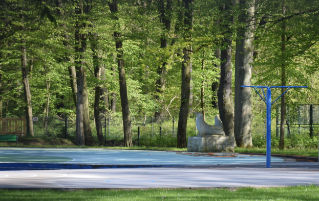 Basketballplatz in einem Park mit Bäumen, einer Bank auf der linken Seite, einem Zaun im Hintergrund und Gras unten.