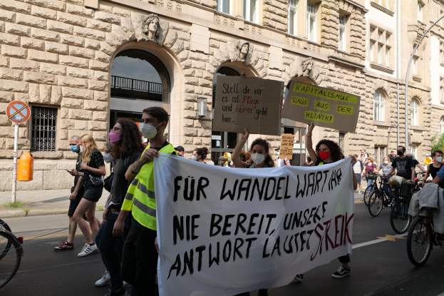 Eine Gruppe von Menschen bei einer Demonstration gegen die Anti-Kriegs-Bewegung, die eine Straße mit Plakaten, Bannern und Fahrrädern entlanggeht, vorbei an einem Gebäude mit architektonischen Details und Bäumen im Hintergrund.