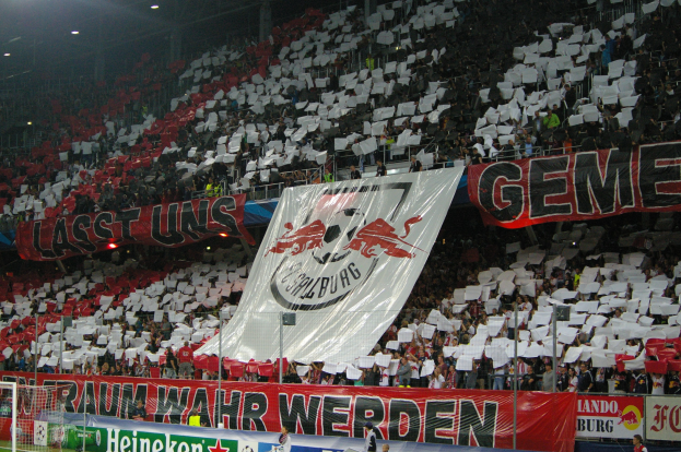 Ein volles Fußballstadion mit Sitz- und Stehplätzen, Bannern, einem Tor und Flutlicht.