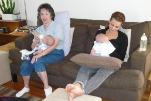 Zwei Frauen sitzen auf einer Couch und halten Babys, mit Kissen drumherum, vor einer weißen Wand, mit einem Schreibtisch, Büchern und einer Zimmerpflanze links daneben.