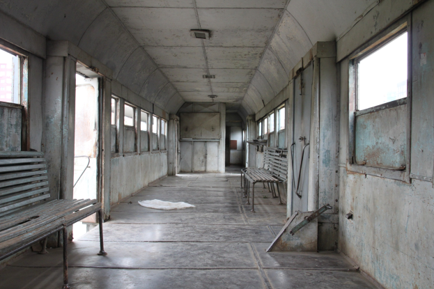 Innensicht eines alten Zugwagens mit hölzernen Bänken, Fenstern auf beiden Seiten und einer Decke, mit einem Gebäude im Freien.