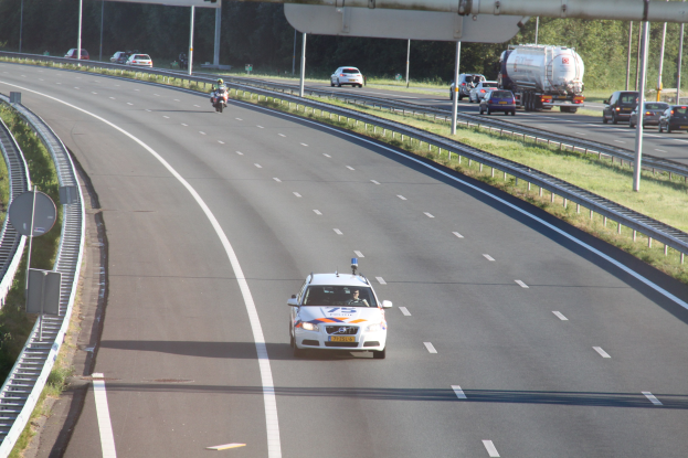 Ein Auto auf einer Autobahn mit einem stationären Polizeiwagen am Rödelfeld, andere Fahrzeuge und Bäume, Geländer, Masten und Planken im Hintergrund.