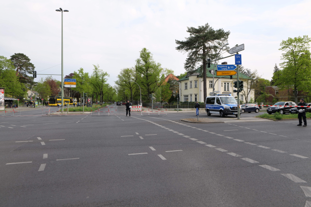 Eine Kreisverkehrausfahrt mit einem parkenden Polizeiwagen, Fahrzeugen auf der Straße, Passanten auf dem Gehweg, Verkehrszeichen, Bäume, Gebäude mit Fenstern und eine bewölkte Himmel.