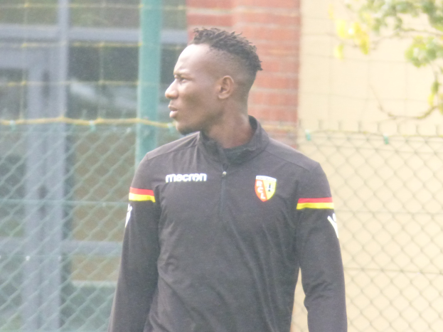 Ein Mann in einer schwarzen Jacke steht auf einem Fussballfeld, umgeben von einem Zaun und einem Baum, und sieht konzentriert aus.