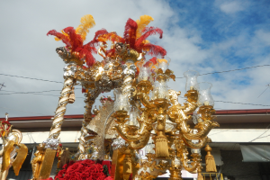 Ein prunkvoller gold- und roter Festwagen mit Blumen und anderen Schmuckelementen in einem Karnevalsumzug, mit einem Gebäude, Strommasten mit Kabeln und einem bewölkten Himmel im Hintergrund.