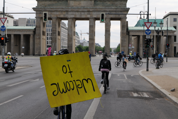 Eine Gruppe von Radfahrern fährt an der Brandenburgertor in Berlin vorbei, einer hält ein gelbes Schild, mit Laternenmasten, Ampeln, Gebäuden, Bäumen und einem klaren Himmel im Hintergrund.