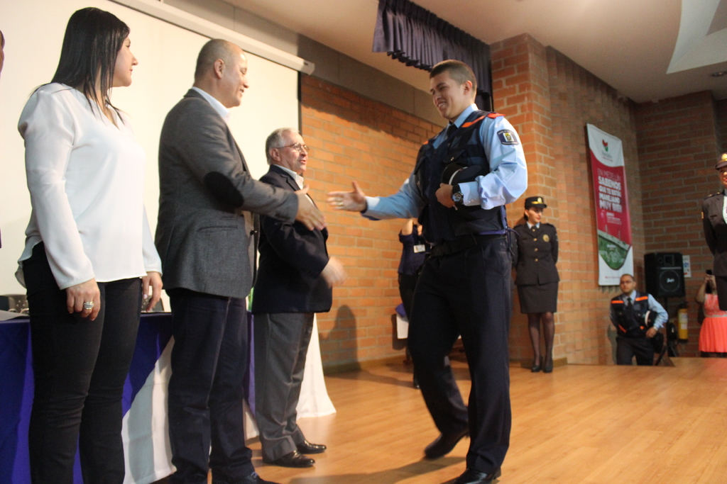 Eine Gruppe von Menschen auf einer Bühne, einer im Uniform mit einer Mütze, eine Frau in einem weißen Hemd, mit einem Banner, Lautsprecher, Vorhängen, Tisch und Wandbildschirm im Hintergrund.