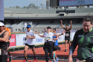 Gruppe von Läufern auf einer Outdoor-Sportanlage in einem Stadion mit Bannern, einer Anzeigetafel, einem Gebäude, Bäumen und einer bewölkten Himmel.